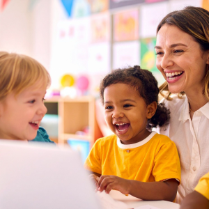 A smiling teacher and three young children are gathered around a laptop in a brightly lit classroom.