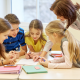 A teacher leans over a table to help a group of students with their work. The students are gathered around a piece of paper, pointing and discussing. There are notebooks and pens on the table.