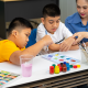 A teacher guides two children, one with a hearing aid, as they paint at a table with watercolours and brushes.